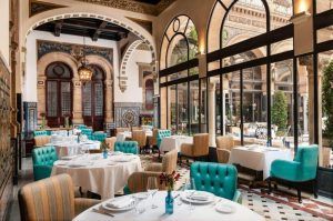Un elegante restaurante refleja el ambiente lujoso del Hotel Alfonso XIII Sevilla con paredes ornamentadas, ventanas arqueadas, sillas turquesas y mesas adornadas con manteles blancos. - ForTwo - Planes para parejas