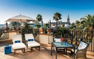 Disfrute de las impresionantes vistas de la ciudad, incluida la torre de la catedral y las palmeras ondulantes, desde la terraza de la azotea del Hotel Alfonso XIII Sevilla, que cuenta con tumbonas y una mesa con sombrilla. - ForTwo - Planes para parejas