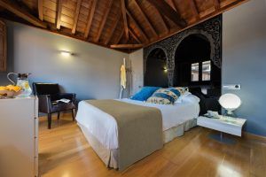 Disfrute de una acogedora habitación con techo de madera, cama con almohadas azules, un acogedor sillón y espejo. La cálida iluminación sobre el suelo de madera evoca el encanto del Hotel Boutique Gar Anat Granada. - ForTwo - Planes para parejas
