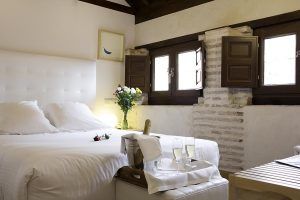 Una acogedora habitación en el Hotel Boutique Gar Anat Granada con una gran cama blanca, flores, champán y pequeñas ventanas con contraventanas de madera oscura. - ForTwo - Planes para parejas