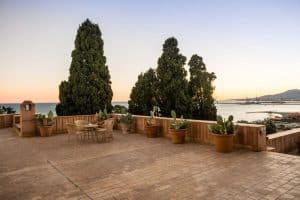 La terraza del Hotel Castillo de Santa Catalina Málaga ofrece un remanso de paz con plantas en macetas y una mesa encantadora, ofreciendo vistas a la costa de Málaga y a las montañas lejanas al atardecer. - ForTwo - Planes para parejas
