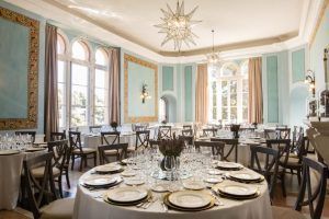 Disfrute de una cena elegante en el Hotel Castillo de Santa Catalina Málaga con mesas redondas, cubiertos, paredes turquesas y grandes ventanales adornados con cortinas beige, todo ello creando un ambiente de inspiración malagueña que complementa maravillosamente las impresionantes vistas del exterior. - ForTwo - Planes para parejas