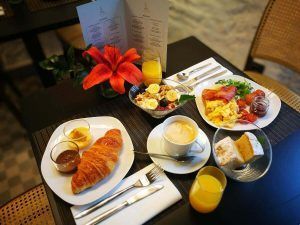El desayuno en el Hotel Legado Alcázar de Sevilla incluye un croissant, café, fruta, huevos, tocino y jugo de naranja bellamente dispuestos alrededor de un centro de mesa con un lirio rojo. - ForTwo - Planes para parejas