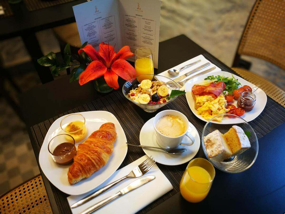 El desayuno en el Hotel Legado Alcázar de Sevilla incluye un croissant, café, fruta, huevos, tocino y jugo de naranja bellamente dispuestos alrededor de un centro de mesa con un lirio rojo. - ForTwo - Planes para parejas