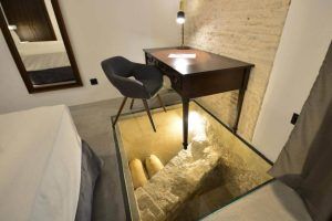 Una habitación del Hotel Legado Alcázar de Sevilla cuenta con un suelo de cristal que muestra ruinas antiguas debajo, y está equipada con un escritorio, una silla y una cama. - ForTwo - Planes para parejas