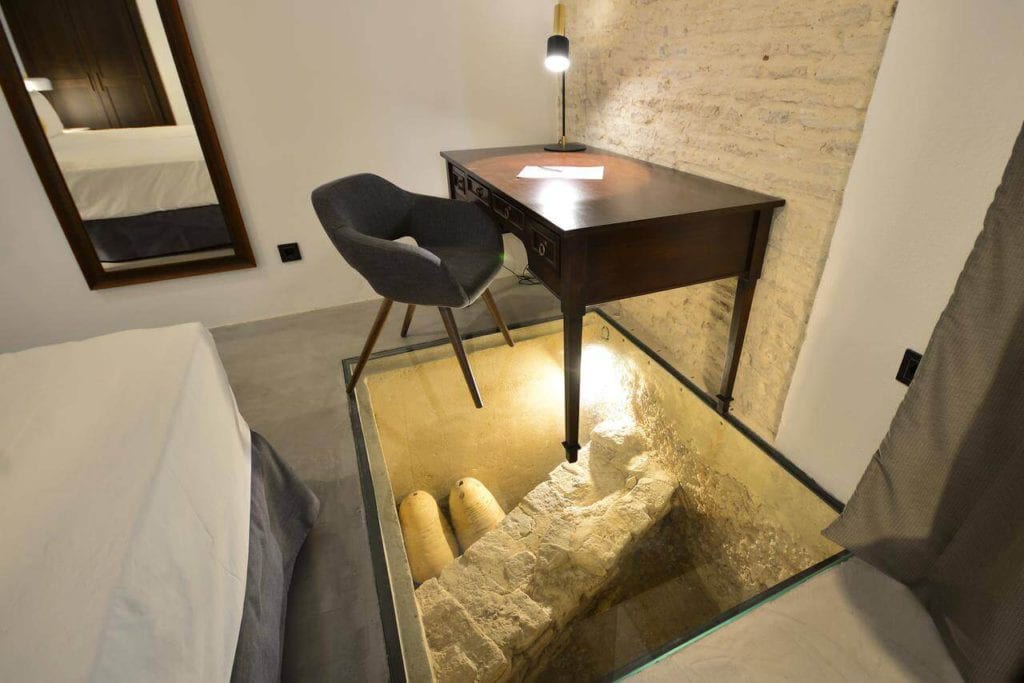Una habitación del Hotel Legado Alcázar de Sevilla cuenta con un suelo de cristal que muestra ruinas antiguas debajo, y está equipada con un escritorio, una silla y una cama. - ForTwo - Planes para parejas