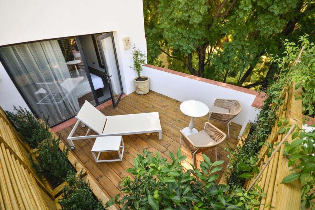 Relájese en el acogedor patio de madera del Hotel Legado Alcázar en Sevilla, que cuenta con sillones, una mesa redonda, plantas exuberantes y vistas a árboles verdes vibrantes. - ForTwo - Planes para parejas