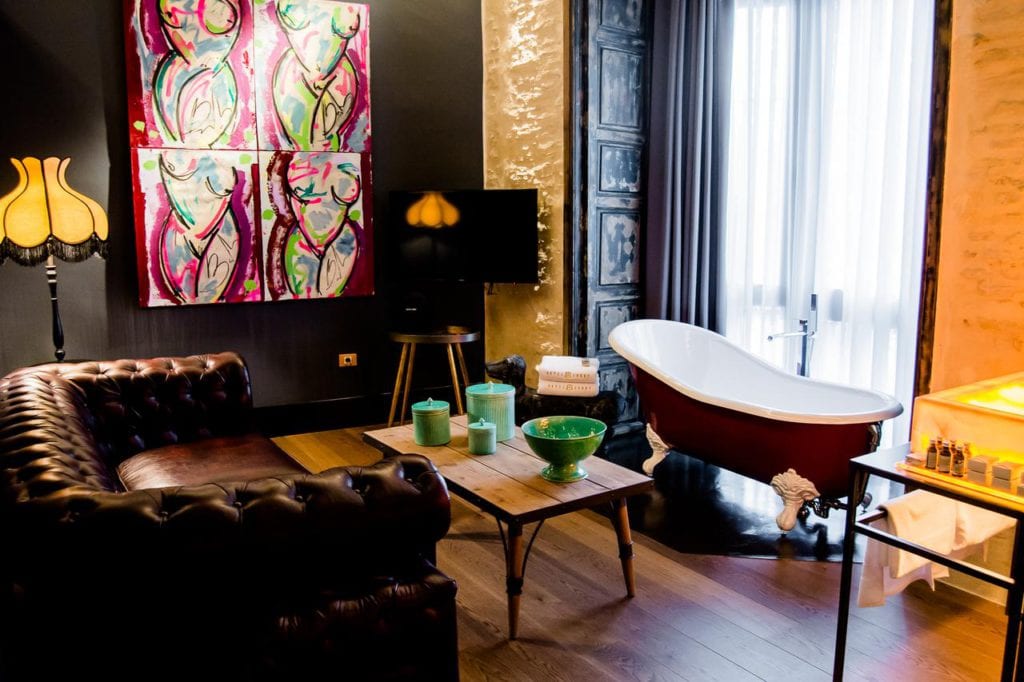 La habitación del vestíbulo del hotel Sevilla ofrece una experiencia elegante con una llamativa bañera con patas de color rojo, un lujoso sofá de cuero, pinturas abstractas y una acogedora lámpara de pie, perfecta para relajarse después de disfrutar de las vibrantes calles de Sevilla. - ForTwo - Planes para parejas