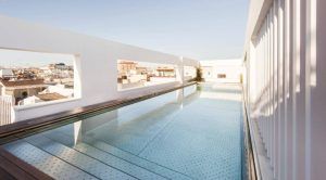 Disfrute de la contemporánea piscina en la azotea del Hotel Mercer (Sevilla), con paredes de cristal transparente que ofrecen impresionantes vistas del horizonte de la ciudad contra un cielo azul claro. - ForTwo - Planes para parejas