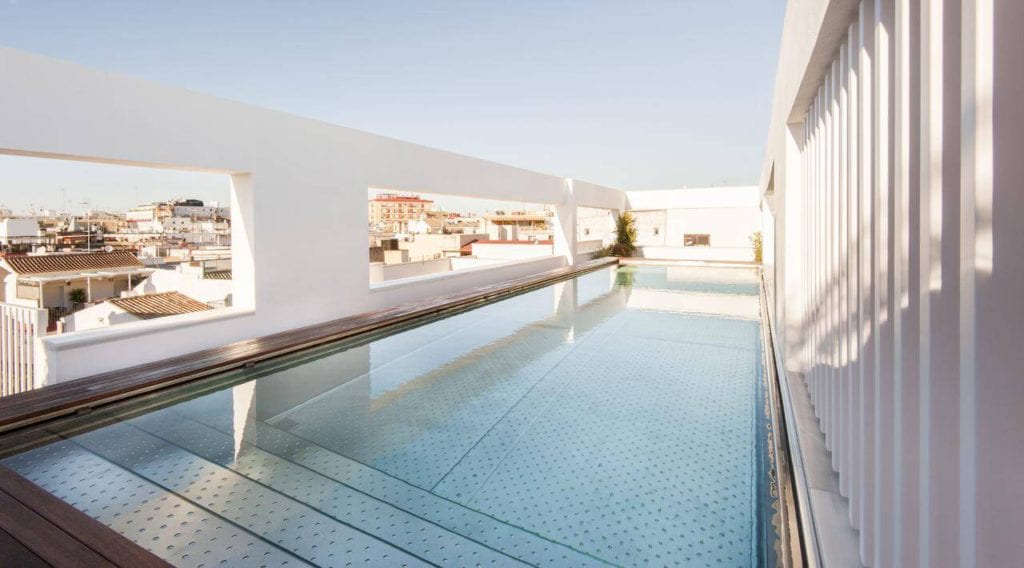 Disfrute de la contemporánea piscina en la azotea del Hotel Mercer (Sevilla), con paredes de cristal transparente que ofrecen impresionantes vistas del horizonte de la ciudad contra un cielo azul claro. - ForTwo - Planes para parejas