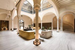 El elegante vestíbulo del Hotel Mercer en Sevilla cuenta con suelos de mármol, techos abovedados, muebles contemporáneos y una columna colocada centralmente bajo un tragaluz. - ForTwo - Planes para parejas