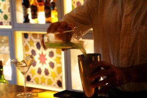 Un barman del Hotel Palacio de Villapanés Sevilla vierte con destreza un líquido verde en una coctelera, enmarcado por unas paredes retroiluminadas de colores vibrantes y una copa de martini esperando. - ForTwo - Planes para parejas