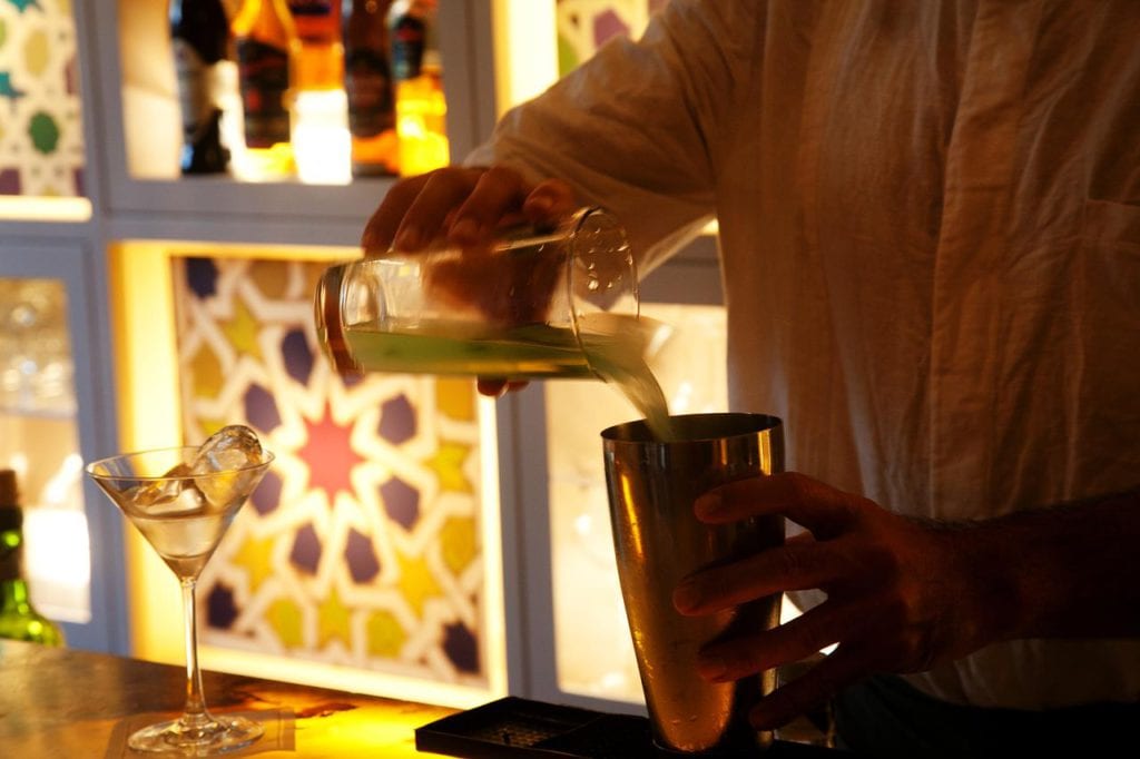 Un barman del Hotel Palacio de Villapanés Sevilla vierte con destreza un líquido verde en una coctelera, enmarcado por unas paredes retroiluminadas de colores vibrantes y una copa de martini esperando. - ForTwo - Planes para parejas