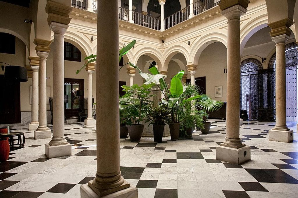 Explore el encanto del Hotel Palacio de Villapanés Sevilla, que muestra un patio histórico adornado con elegantes arcos, columnas, un clásico piso a cuadros y vibrantes plantas en macetas. - ForTwo - Planes para parejas