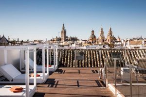 Disfrute del salón en la azotea del Hotel Palacio de Villapanés Sevilla, equipado con tumbonas e impresionantes vistas del paisaje urbano, resaltado por torres históricas enmarcadas por un cielo azul claro. - ForTwo - Planes para parejas