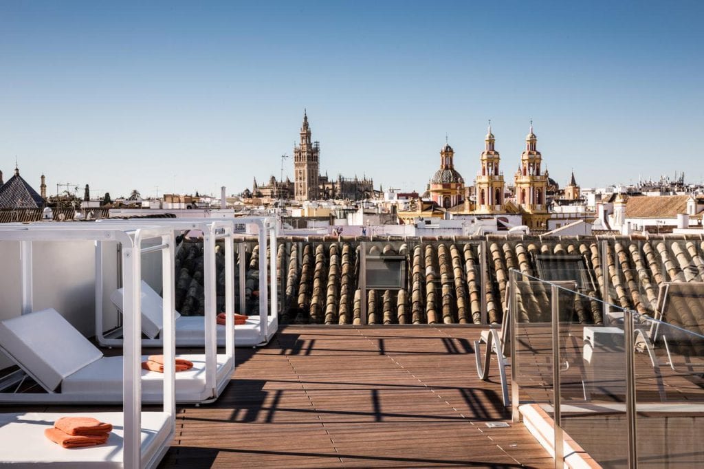 Disfrute del salón en la azotea del Hotel Palacio de Villapanés Sevilla, equipado con tumbonas e impresionantes vistas del paisaje urbano, resaltado por torres históricas enmarcadas por un cielo azul claro. - ForTwo - Planes para parejas