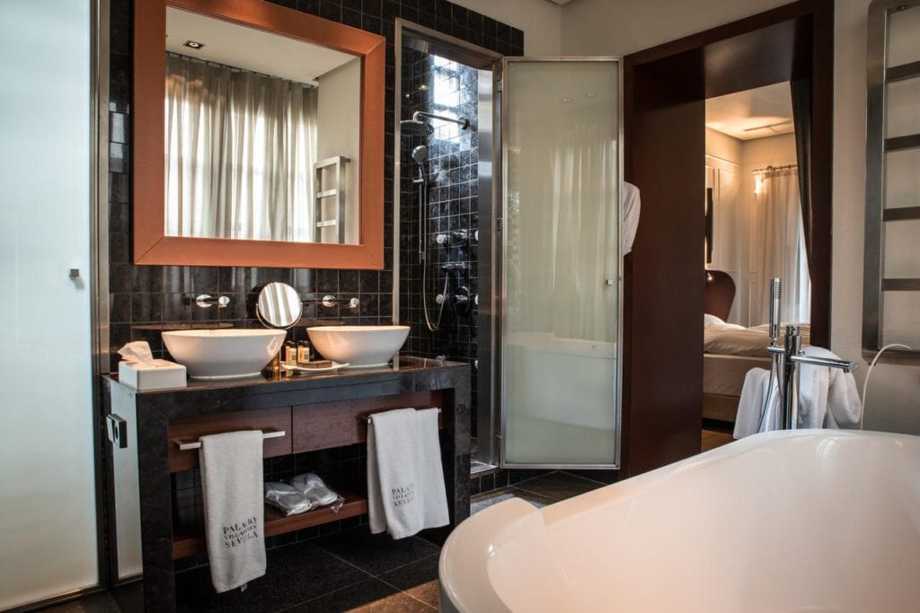 El moderno baño del Hotel Palacio de Villapanés Sevilla incluye lavabos dobles, ducha de cristal y bañera. Suaves toallas cuelgan cuidadosamente bajo el tocador, ofreciendo vistas al elegante dormitorio. - ForTwo - Planes para parejas