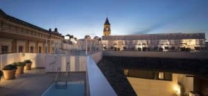 La azotea del Hotel Posada del Lucero Sevilla ofrece una piscina serena, macetas de terracota y vistas panorámicas de los edificios circundantes con una torre de reloj a lo lejos bellamente recortada al anochecer. - ForTwo - Planes para parejas