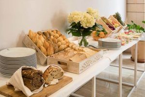 En el Hotel Rey Alfonso X Sevilla se dispone una mesa buffet con surtido de panes, frutas frescas, platos elegantes y un hermoso arreglo floral. - ForTwo - Planes para parejas