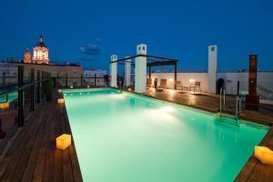 Al anochecer, la piscina de la azotea del Hotel Vincci Selección Posada del Patio Málaga ofrece una iluminación ambiental sobre una terraza de madera con vistas lejanas a los edificios históricos iluminados de Málaga, creando un entorno perfecto para los huéspedes. - ForTwo - Planes para parejas