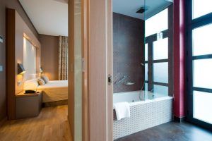 Experimente el encanto contemporáneo de una habitación del Hotel Vincci Selección Posada del Patio Málaga, equipada con una cómoda cama, lámparas de noche y un baño abierto con una elegante mampara de cristal y bañera. - ForTwo - Planes para parejas
