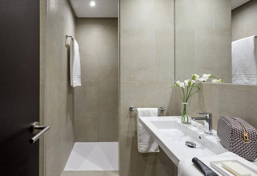 Este baño minimalista, que refleja el estilo elegante del Hotel Posada del Lucero Sevilla, incluye azulejos beige, un lavabo blanco, un toallero, un jarrón de flores y un bolso estampado en el mostrador. - ForTwo - Planes para parejas