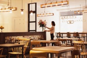En la acogedora cafetería del Hotel 7 Islas Madrid, una mujer arregla elegantemente flores entre muebles de madera y una iluminación cálida, creando un oasis de serenidad en el centro de Madrid. - ForTwo - Planes para parejas