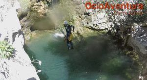 Una persona que lleva un traje de neopreno salta a una piscina rocosa de color turquesa en medio de una exuberante vegetación en Jorox, mostrando "Barranquismo en Jorox de la mano de Ocio Aventura (Málaga)" arriba. - ForTwo - Planes para parejas