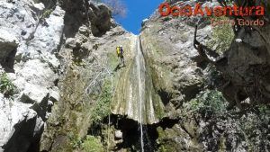 Vestido con equipo amarillo, una persona desciende en rápel por una cascada rocosa y musgosa bajo el cielo azul claro en Jorox, encarnando la emoción y el espíritu del "Barranquismo en Jorox de la mano de Ocio Aventura" (Málaga). - ForTwo - Planes para parejas