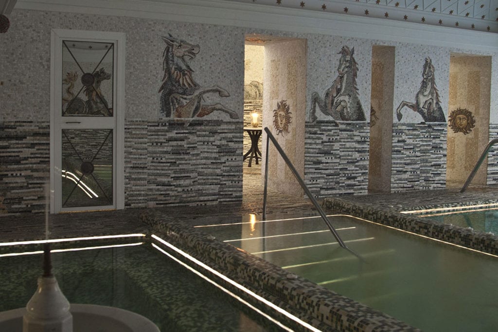 El Circuito Termal con Masaje de Termas de Hispalis en Sevilla ofrece un oasis de serenidad con paredes de mosaico de caballos y soles, iluminación ambiental que se refleja en la superficie del agua y espacios acogedores para relajarse y descansar. - ForTwo - Planes para parejas