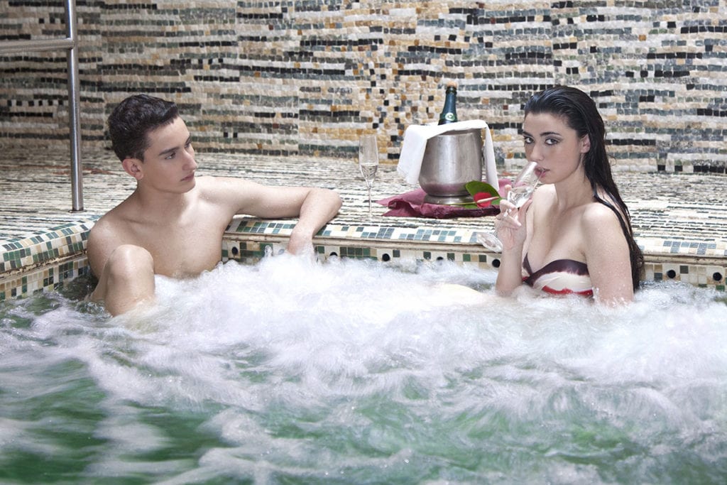 Dos personas se relajan en un jacuzzi; una bebe champán mientras una botella reposa sobre hielo, muy parecido al ambiente relajante del Circuito Termal con Masaje en Termas de Hispalis, Sevilla. - ForTwo - Planes para parejas
