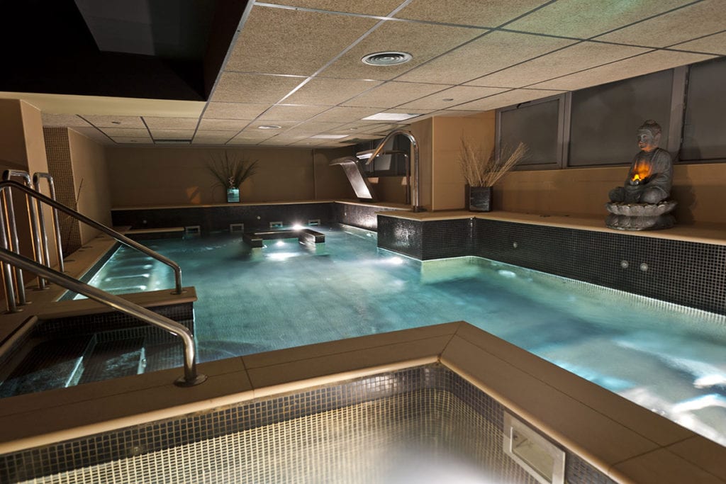 Experimente la serenidad con el "Circuito spa con Masaje e infusión o zumo en Kumaras Spa (Umbrete)", donde una iluminación suave, pasamanos de metal y una estatua decorativa de Buda crean un ambiente tranquilo alrededor de la tranquila piscina interior del spa, invitándolo a relajarse y disfrutar de momentos relajantes. - ForTwo - Planes para parejas