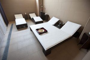 La moderna sala de spa del Kumaras Spa (Umbrete) cuenta con cuatro sillones con cojines y una bandeja decorativa sobre la mesa, ideal para relajarse después del masaje. Las paredes y azulejos neutros realzan la atmósfera relajante de su experiencia en el Circuito spa con Masaje e Infusión o Zumo. - ForTwo - Planes para parejas