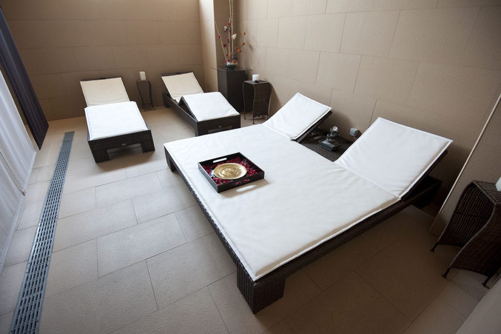 La moderna sala de spa del Kumaras Spa (Umbrete) cuenta con cuatro sillones con cojines y una bandeja decorativa sobre la mesa, ideal para relajarse después del masaje. Las paredes y azulejos neutros realzan la atmósfera relajante de su experiencia en el Circuito spa con Masaje e Infusión o Zumo. - ForTwo - Planes para parejas
