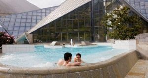 Una pareja se relaja en una piscina al aire libre junto al edificio triangular de cristal de Entrada al spa de Caldea, un famoso spa en Andorra, rodeado de impresionantes árboles y montañas. - ForTwo - Planes para parejas