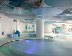 Descubra una elegante piscina cubierta en Caldea, Andorra, con paneles circulares en el techo e iluminación ambiental. El texto de Likids en la pared añade un toque de estilo a su experiencia en la Entrada al spa de Caldea. - ForTwo - Planes para parejas