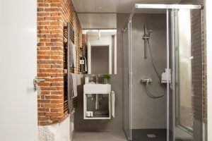 Baño moderno inspirado en los Apartamentos Boutique Eric Vökel, con pared de ladrillo, ducha de cristal, lavabo blanco compacto y azulejos grises. Las estanterías exhiben toallas y artículos de aseo, capturando la elegancia sobria típica de los apartamentos boutique. - ForTwo - Planes para parejas