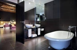Un moderno baño con azulejos negros, lavabos dobles, un gran espejo y una bañera exenta destila elegancia de estilo boutique. El dormitorio, al fondo, luce el sofisticado diseño inspirado en el Hotel Eurostars Sevilla Boutique. - ForTwo - Planes para parejas