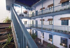 En el Hotel Hospes Las Casas del Rey de Baeza en Sevilla, un patio de estilo colonial cuenta con barandillas azules, plantas vibrantes y dos pisos de habitaciones bellamente iluminadas, que fomentan una atmósfera nocturna serena. - ForTwo - Planes para parejas
