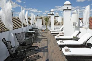 Relájese en la terraza de la azotea del Hotel Hospes Las Casas del Rey de Baeza de Sevilla, que cuenta con tumbonas, muebles de mimbre y cortinas blancas que ofrecen un impresionante refugio frente a un techo de tejas y un cielo azul intenso. - ForTwo - Planes para parejas