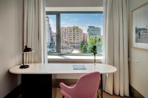 La habitación del Hotel Barceló Torre de Madrid cuenta con una acogedora silla rosa y un escritorio junto a la ventana, ideal para disfrutar de las impresionantes vistas de la ciudad de Madrid. - ForTwo - Planes para parejas