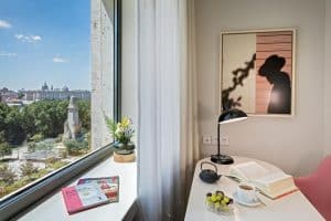 Un escritorio iluminado por el sol, con libros, café, uvas y una lámpara, se encuentra junto a una ventana con vistas al Hotel Barceló Torre de Madrid y al parque cercano. La silueta de un sombrero adorna la pared. - ForTwo - Planes para parejas