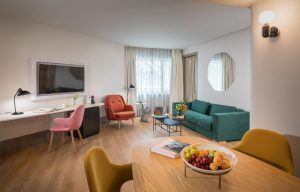 Descubra la elegancia moderna en el salón del Hotel Barceló Torre de Madrid, con un sofá verde azulado y un sillón rojo. Relájese junto a la mesa redonda de madera con un frutero, mientras disfruta del televisor de pared. Experimente la hospitalidad madrileña en este ambiente inspirado en un hotel. - ForTwo - Planes para parejas