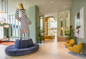 El moderno lobby del Hotel Barceló Torre de Madrid luce una escultura de rayas sobre un sofá circular, con paredes verdes y sillas amarillas, creando un diseño elegante y espacioso. - ForTwo - Planes para parejas