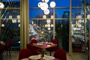 Ubicado en el Hotel Barceló Torre de Madrid, el interior del restaurante cuenta con sillas rojas, iluminación ornamentada y grandes ventanales que ofrecen vistas panorámicas de la bulliciosa calle de la ciudad por la noche. - ForTwo - Planes para parejas