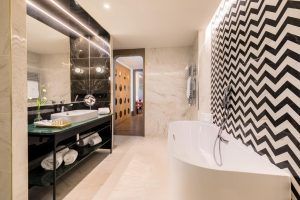 El baño del Hotel Barceló Torre de Madrid luce una pared con detalles en chevron en blanco y negro, una bañera independiente y espejos grandes, fusionando el lujo moderno con el estilo característico del hotel. - ForTwo - Planes para parejas