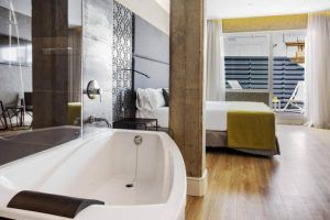 Alójese en el Hotel Catalonia Giralda de Sevilla para disfrutar de una habitación contemporánea con bañera, suelo de madera, cama doble y puertas de cristal que dan a un balcón iluminado por el sol. - ForTwo - Planes para parejas