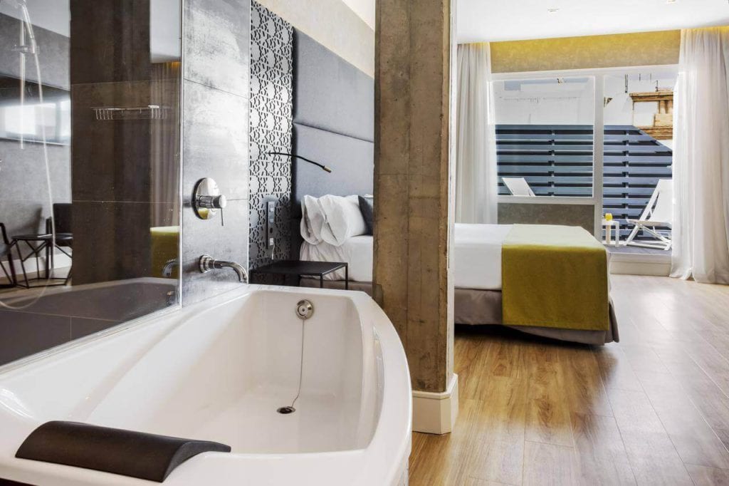 Alójese en el Hotel Catalonia Giralda de Sevilla para disfrutar de una habitación contemporánea con bañera, suelo de madera, cama doble y puertas de cristal que dan a un balcón iluminado por el sol. - ForTwo - Planes para parejas