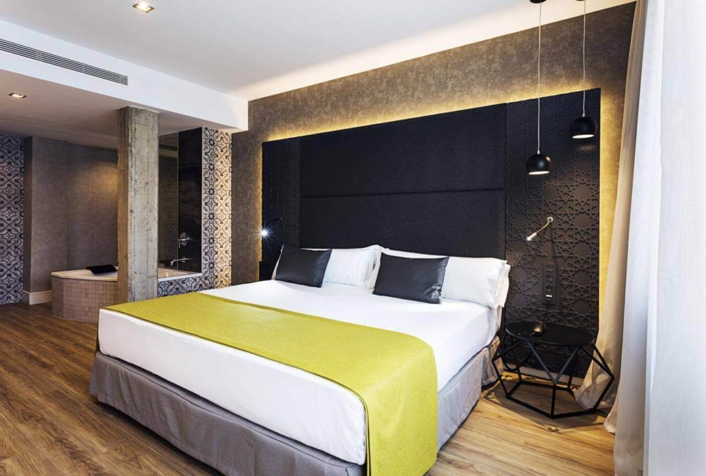 Descubra la elegancia contemporánea en el Hotel Catalonia Giralda con una iluminación elegante, una cama espaciosa, una acogedora manta amarilla y cálidos pisos de madera. - ForTwo - Planes para parejas