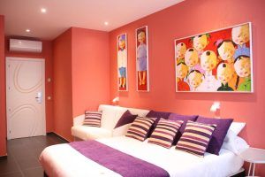 Una acogedora habitación del Hotel Indiana (Madrid) cuenta con paredes rojas, arte vibrante, un sofá blanco y una cama con almohadas y mantas de rayas moradas. - ForTwo - Planes para parejas